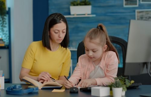 Schoolgirl writing on notebook and checking book for information while mother helping with remote education. Parent and young child doing homework on textbook at desk for online classes.